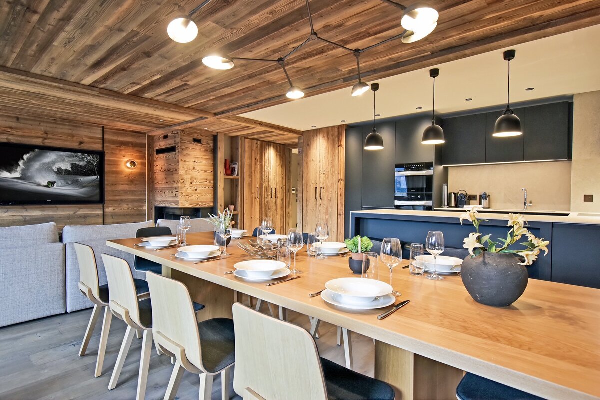 Elegant alpine chalet dining room featuring a long wooden table set for dinner, wooden walls, and a modern open kitchen.