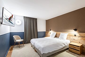 Modern, cozy bedroom with twin beds, wooden headboard, brown/blue walls, TV showing skiing, and a light chair. Carpeted floor.