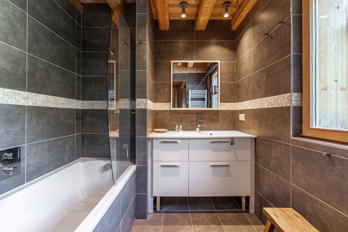 A modern bathroom featuring dark grey tiles with a decorative pebble band, a bathtub, vanity, mirror, and wooden ceiling beams.