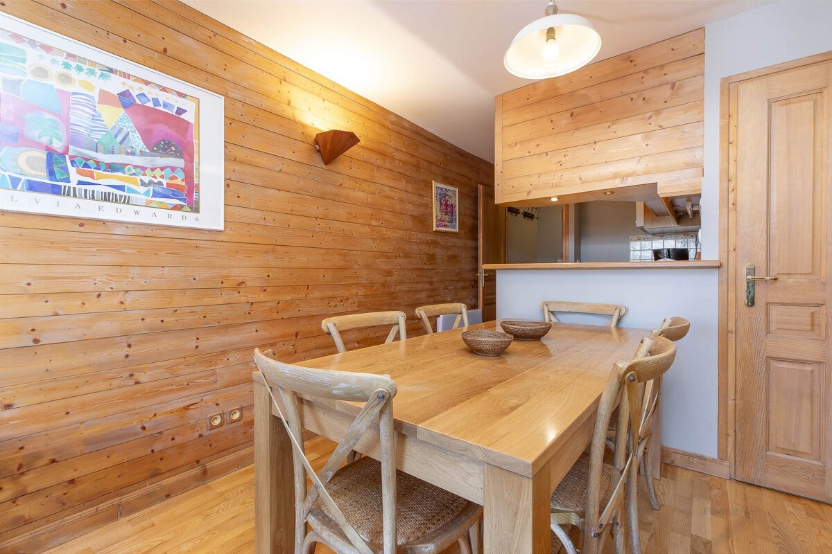 A rustic dining area featuring a wooden table and chairs, wood-paneled walls, colorful artwork, and a kitchen pass-through.