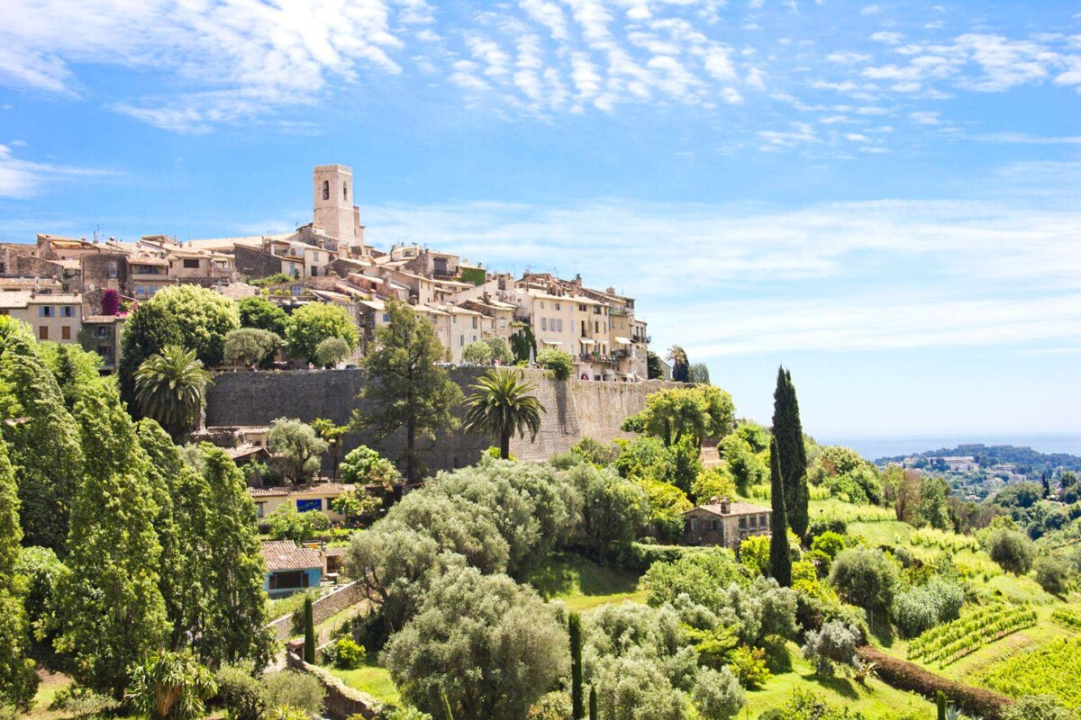 A picturesque hilltop village with a prominent tower, surrounded by lush green trees and vineyards, under a bright blue sky. The sea is visible in the distance.