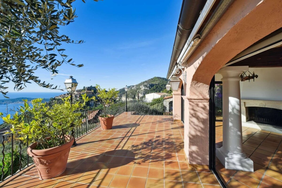 Sunny terrace with terracotta tiles, potted plants, and pink arches overlooking a blue sea, coastal town, and green mountains.