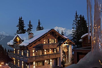 Warmly lit wooden chalet covered in snow at dusk, with towering mountains and icicles, evoking a cozy winter retreat.