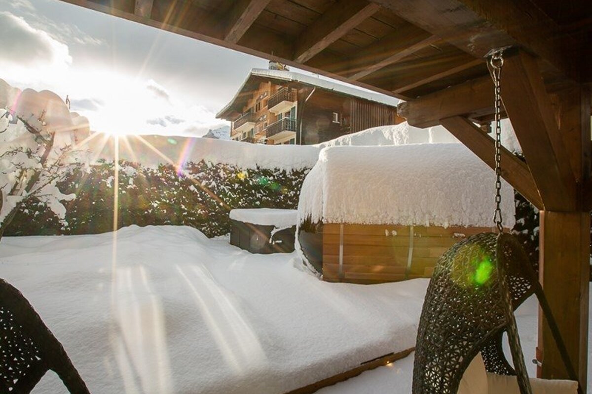 Bright winter sun shines over a snow-covered patio featuring a barrel sauna, viewed from a wooden porch.