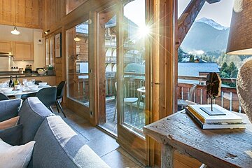 Sunlight streams into a cozy wooden chalet interior with a balcony overlooking scenic, snow-capped mountains.