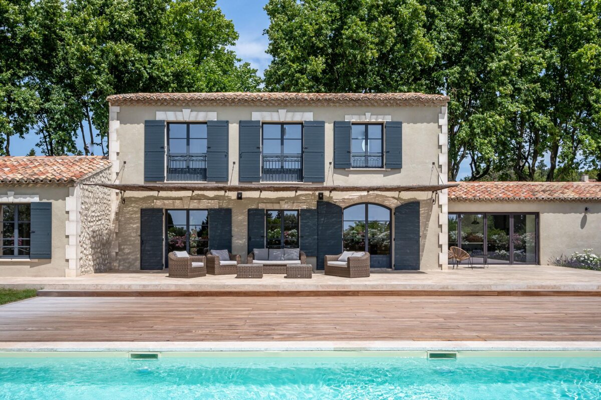 An elegant stone house with dark shutters, a spacious patio and deck, and a sparkling blue swimming pool. Lush green trees frame the property.