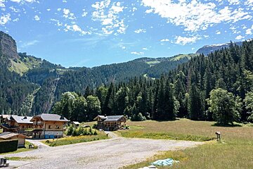 A sunny mountain landscape featuring wooden chalets, dense green forests, and a ski lift under a blue sky with fluffy white clouds.