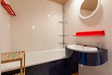 A bathroom with white tiled walls, a bathtub, a unique blue wall-mounted sink, a round mirror, and red accent shelves/hangers.