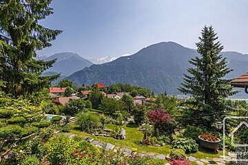 A lush garden overlooks a village nestled in a mountain valley, with snow-capped peaks in the distance and a paraglider under a clear sky.