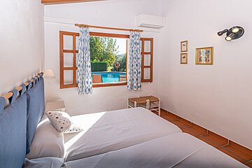 A bright bedroom with twin beds and white walls overlooks a pool through a wooden window. Blue patterned curtains, an AC unit, and wall art complete the space.