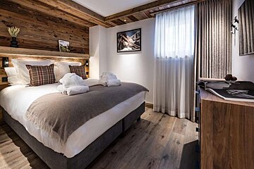 Cozy, rustic bedroom featuring a large bed with plaid pillows, white towels, and a wooden accent wall/ceiling. Hardwood floors and sheer curtains complete the look.