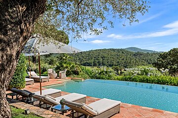 A luxurious infinity pool with lounge chairs on a terracotta patio, shaded by an old tree, overlooks lush green hills under a bright blue sky.