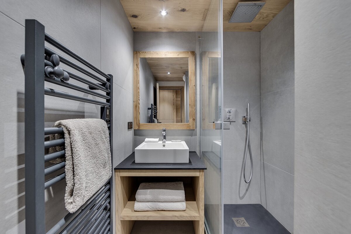 A modern bathroom with grey tiled walls, a wooden ceiling and vanity, a vessel sink, a walk-in shower with glass, and a black towel warmer.