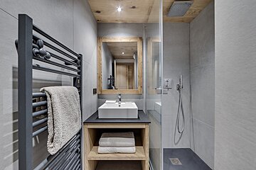 A modern bathroom with grey tiled walls, a wooden ceiling and vanity, a vessel sink, a walk-in shower with glass, and a black towel warmer.