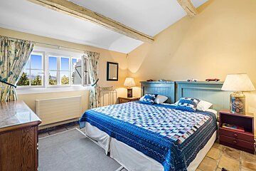 A rustic bedroom featuring a blue quilt-covered bed, yellow walls, exposed ceiling beams, and a window looking out on a sunny day.