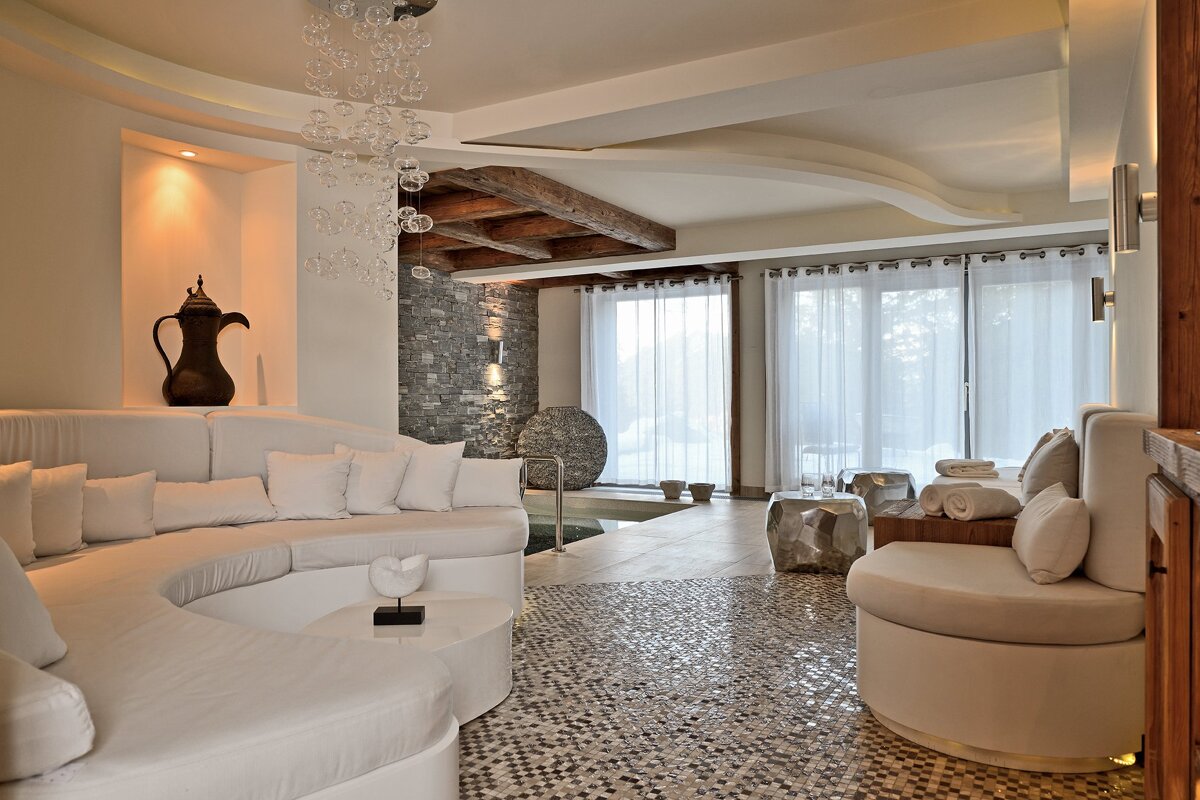 Luxurious spa lounge featuring curved white seating, rustic wood ceiling beams, a stone accent wall, and a mosaic floor.