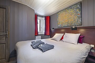 Modern bedroom with grey paneled walls, a large bed, abstract art, and red curtains. Folded towels await on the bed.