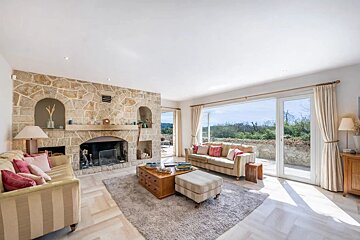 A spacious, bright living room features a large stone fireplace, two cozy sofas, and expansive glass doors opening to a sunny outdoor view.