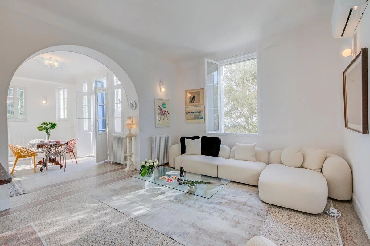 A bright, white living room with a curved sofa, glass coffee table, and an archway leading to a dining area.