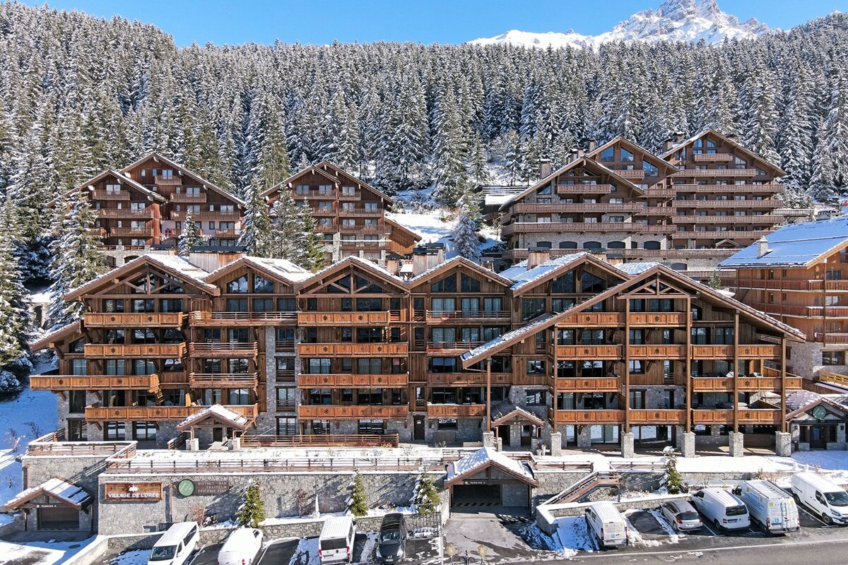 Large wooden chalets at a ski resort, nestled against a snow-covered pine forest under a clear blue sky.