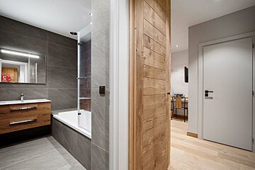 Modern bathroom with grey tiles and wooden vanity, next to a hallway with a sliding wooden door and light wood floors.