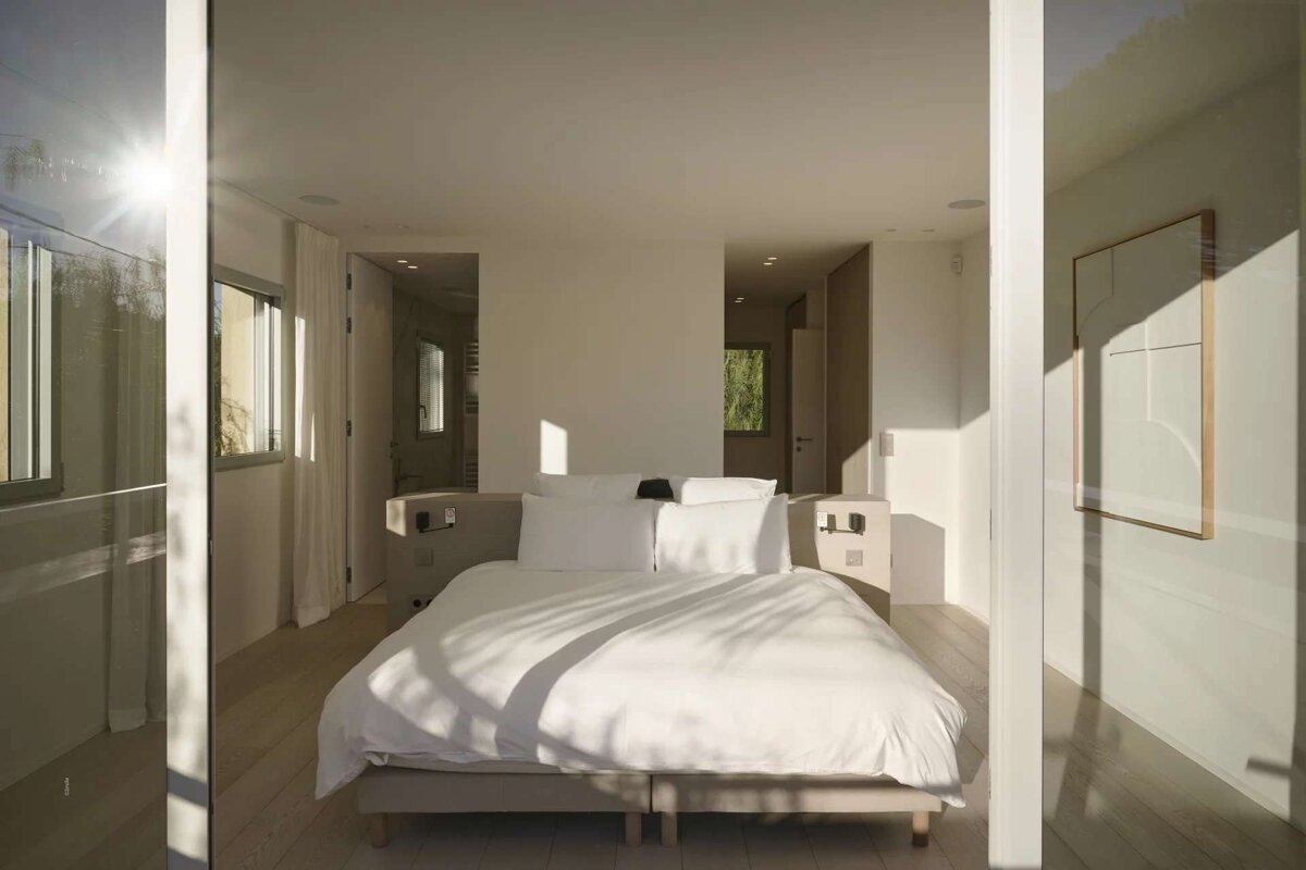 A bright, minimalist bedroom with a large white bed, bathed in natural sunlight creating sharp shadows and a serene atmosphere.