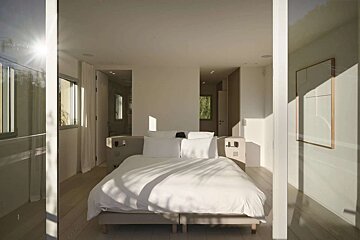 A bright, minimalist bedroom with a large white bed, bathed in natural sunlight creating sharp shadows and a serene atmosphere.