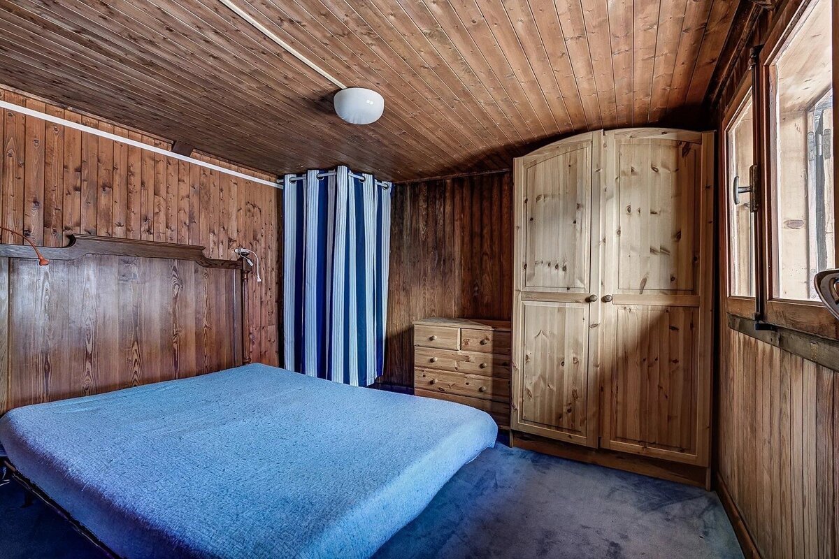 A wood-paneled bedroom features a blue bed, wooden wardrobe, dresser, and striped curtains. A window and blue carpet complete the rustic setting.