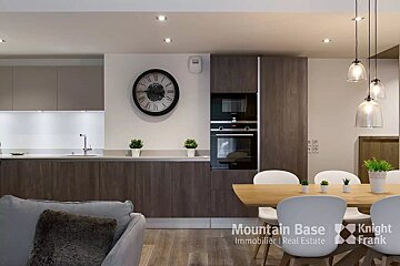 Modern open-plan kitchen and dining area with dark wood cabinetry, integrated appliances, a large wall clock, and a light wood dining table with white chairs.
