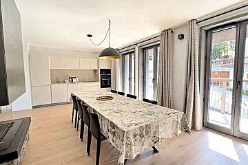 A modern open-plan kitchen and dining area featuring a long table with chairs, minimalist cabinets, and large glass doors with checkered curtains.