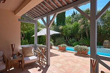 A relaxing covered patio with outdoor furniture, terracotta tiles, overlooking a lush garden with diverse plants and a sparkling blue pool.