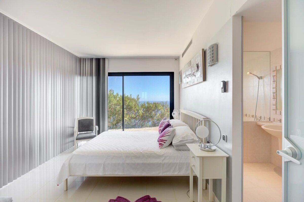 Modern bedroom with a large window overlooking the ocean, a white bed, and an open door revealing an ensuite bathroom.