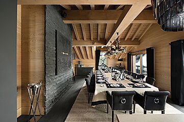 Elegant dining room with a long table, dark chairs, light wood walls/ceiling, exposed beams, and a prominent dark stone feature wall.