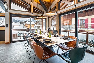 Modern chalet dining room with a set table, stylish leather chairs, and large windows overlooking a snowy alpine village.
