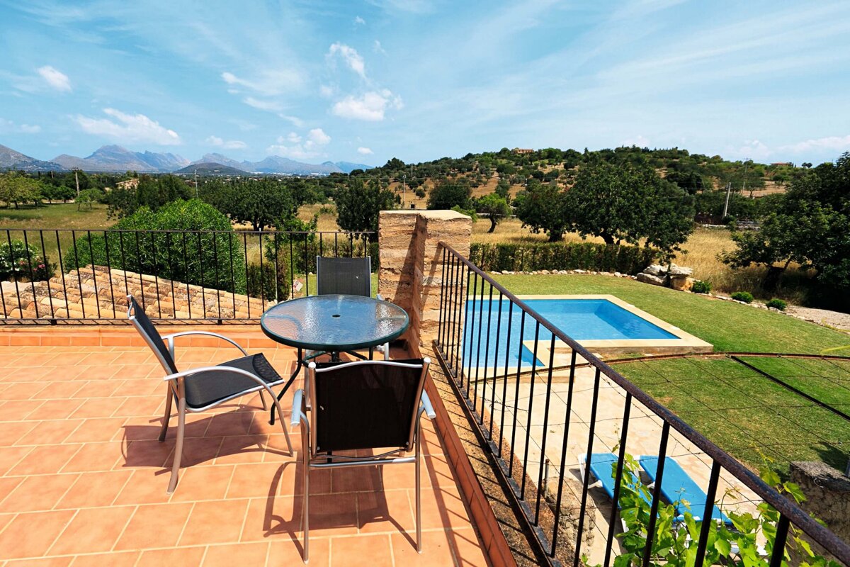 A sunny terracotta terrace with a table and chairs overlooks a vibrant green landscape, distant mountains, and a refreshing blue swimming pool.