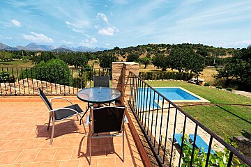 A sunny terracotta terrace with a table and chairs overlooks a vibrant green landscape, distant mountains, and a refreshing blue swimming pool.