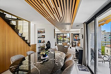 Modern apartment interior with a black marble dining table, slatted wood ceiling, a wooden staircase, and large sliding doors to a balcony.