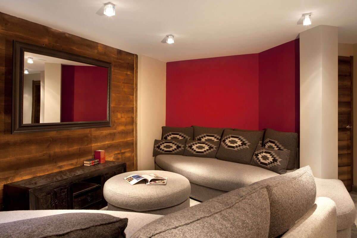 A cozy living area with a bright red accent wall, rustic wood paneling, a large gray sectional sofa with patterned pillows, and a round ottoman.