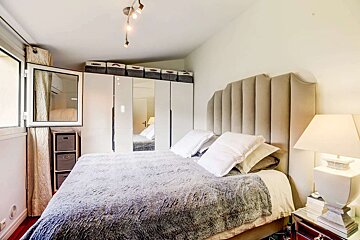 A cozy bedroom featuring a large bed with a beige headboard, grey bedspread, mirrored wardrobe, and a window, illuminated by modern track lighting.