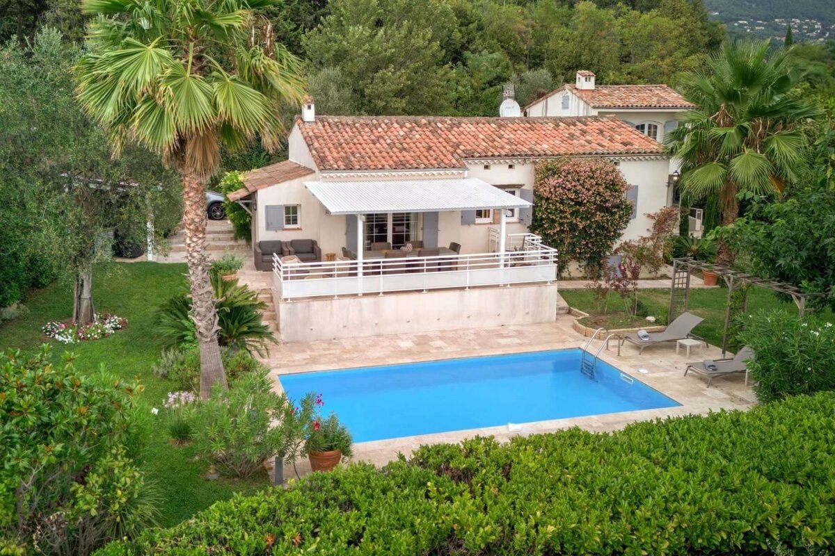 An aerial view shows a charming house with a terracotta roof and a large balcony, overlooking a bright blue swimming pool, all nestled among lush trees and greenery.