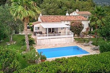 An aerial view of a charming white house with a red roof, large patio, and bright blue swimming pool, surrounded by lush green Mediterranean gardens.
