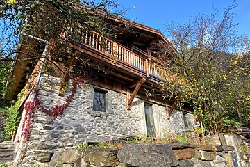 A rustic stone house with a wooden upper floor and balcony, adorned with red ivy and autumn leaves, under a bright blue sky.