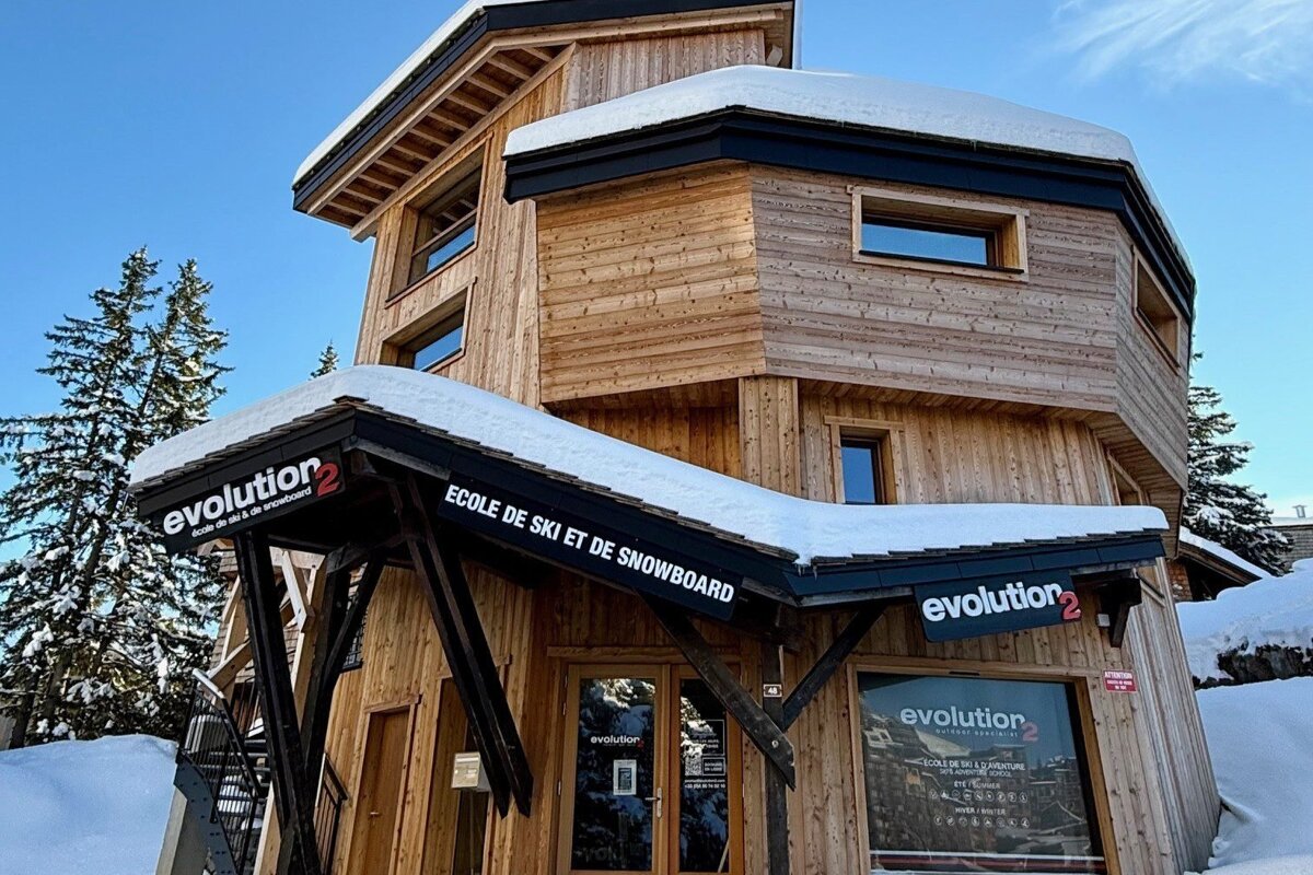 A snow-covered wooden Evolution 2 ski and snowboard school building with trees and blue sky, surrounded by snow.