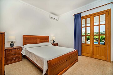 Bright bedroom with a wooden bed, white walls, tiled floor, and French doors opening to an outdoor patio with a pool and greenery.