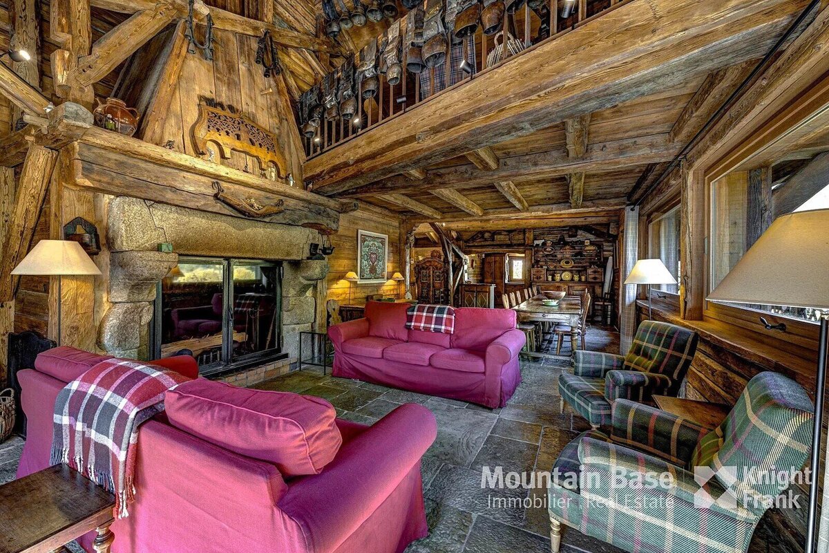 Rustic chalet living room with exposed wooden beams, a stone fireplace, red sofas, plaid armchairs, and a dining area. Cowbells adorn an upper balcony.
