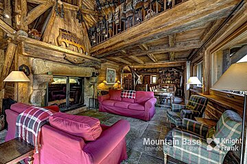 Rustic chalet living room with exposed wooden beams, a stone fireplace, red sofas, plaid armchairs, and a dining area. Cowbells adorn an upper balcony.