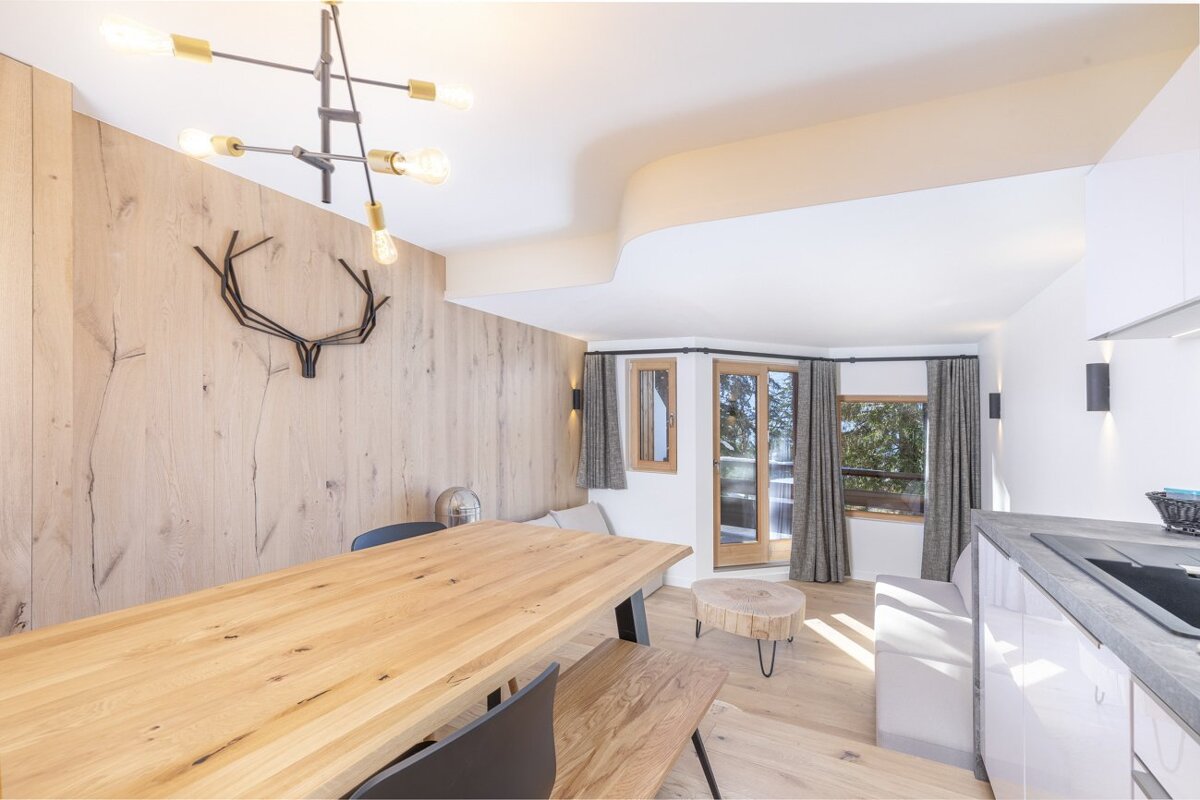 Bright, modern open-plan room featuring a light wood dining area, wooden wall with deer antlers, kitchen, and large windows with outdoor views.