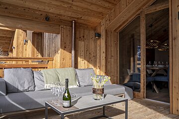 Cozy wooden chalet balcony with a grey sofa, champagne, and flowers on a coffee table. An indoor dining area is visible through glass doors.