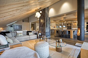 Cozy, modern chalet interior with a slanted wood ceiling, a large grey sectional, a glass coffee table, and a sleek cylindrical fireplace.
