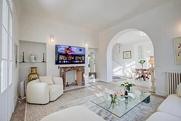 Bright white living room featuring a TV above a marble fireplace, modern white sofas, and a glass coffee table, with an archway leading to a dining area.